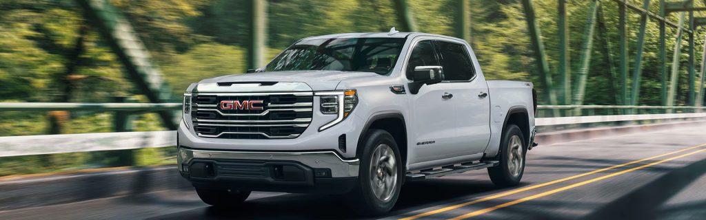 White GMC Sierra pickup truck driving across a bridge on a scenic road surrounded by green trees and motion blur in the background.