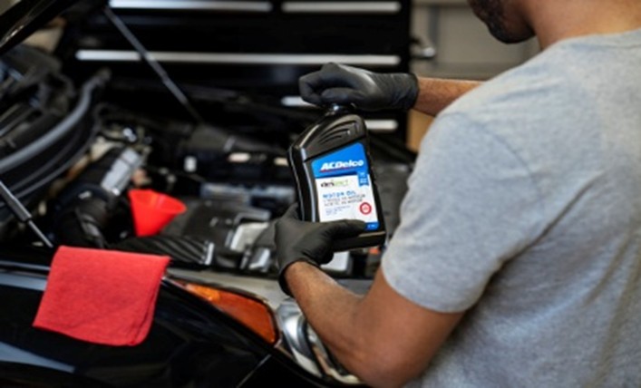 Technician holding engine oil beside an open vehicle hood during a routine maintenance service.